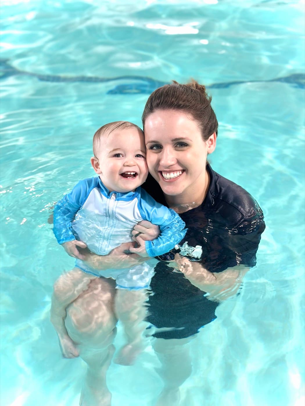 Instructor Katrina smiling in the pool while holding a happy baby in a swim shirt